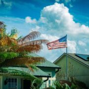 American flag waving on a flagpole in front of a house with palm trees and a blue sky.