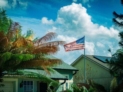 American flag waving on a flagpole in front of a house with palm trees and a blue sky.