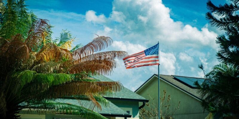 American flag waving on a flagpole in front of a house with palm trees and a blue sky.