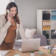 Professional woman standing at a desk, talking on a cellphone, with a laptop and office supplies in a bright office environment.