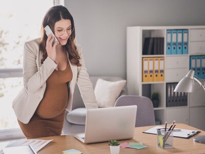Professional woman standing at a desk, talking on a cellphone, with a laptop and office supplies in a bright office environment.