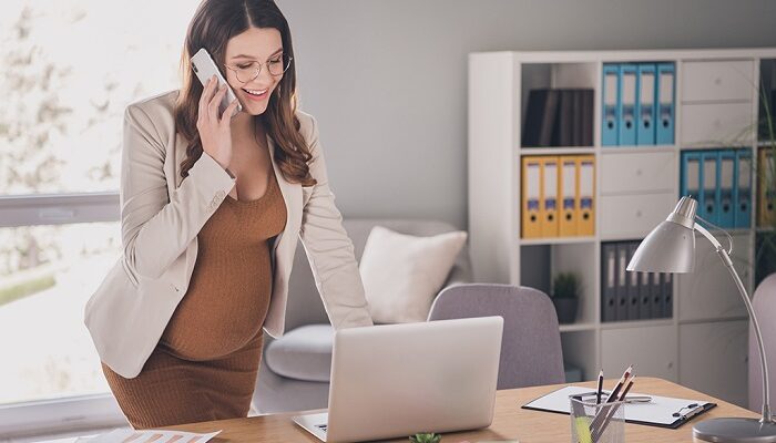 Professional woman standing at a desk, talking on a cellphone, with a laptop and office supplies in a bright office environment.