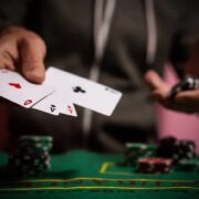 A poker player extends two playing cards toward the camera at a green felt table with chip stacks in the background.