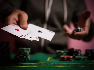 A poker player extends two playing cards toward the camera at a green felt table with chip stacks in the background.