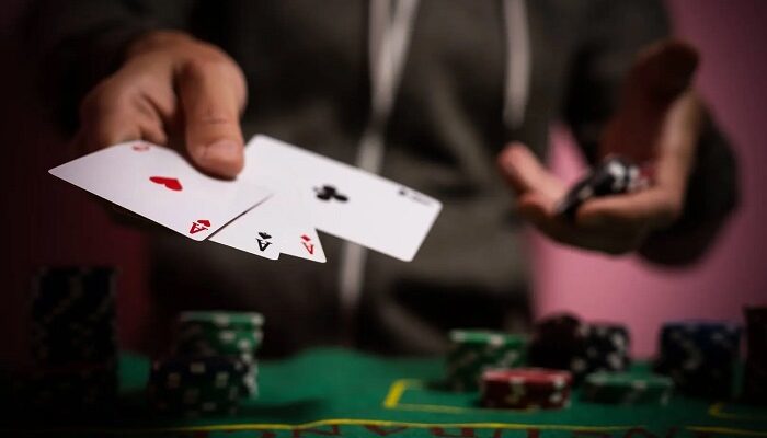 A poker player extends two playing cards toward the camera at a green felt table with chip stacks in the background.