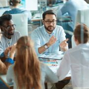 Group of coworkers around a table in a glass-walled office, discussing charts and ideas during a meeting.