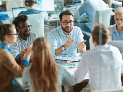 Group of coworkers around a table in a glass-walled office, discussing charts and ideas during a meeting.