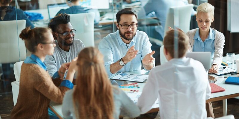 Group of coworkers around a table in a glass-walled office, discussing charts and ideas during a meeting.