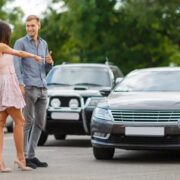 Man and woman stand in a parking lot beside a brown sedan, smiling and chatting with each other.