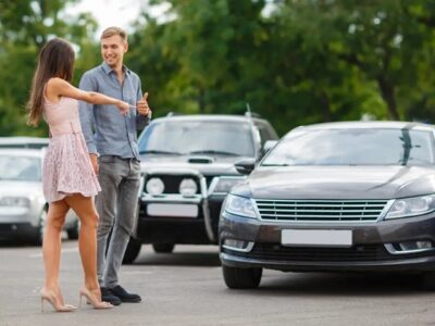 Man and woman stand in a parking lot beside a brown sedan, smiling and chatting with each other.