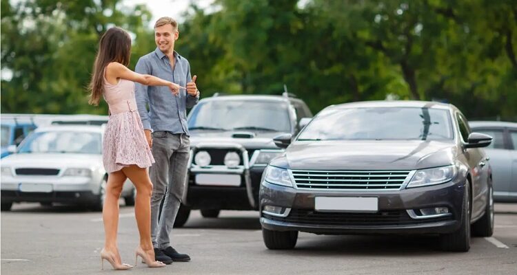 Man and woman stand in a parking lot beside a brown sedan, smiling and chatting with each other.