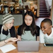 Three young women in aprons gather around a laptop in a café, looking intently at the screen as they discuss a project around the table.