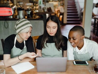 Three young women in aprons gather around a laptop in a café, looking intently at the screen as they discuss a project around the table.