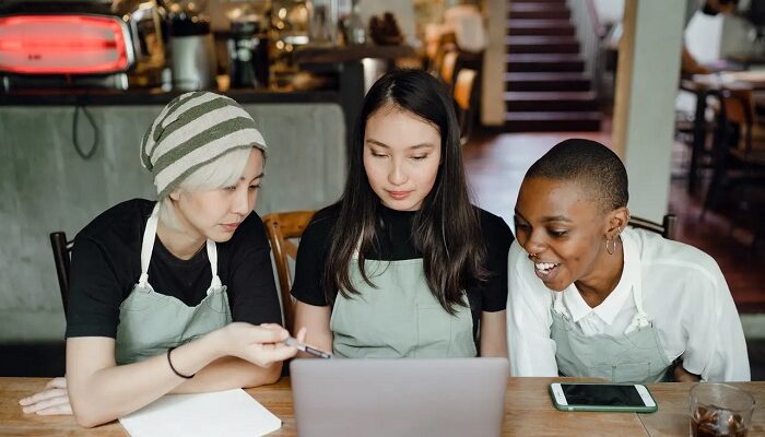 Three young women in aprons gather around a laptop in a café, looking intently at the screen as they discuss a project around the table.