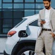 Businessman in a light suit stands beside a white electric SUV, looking at his phone as the charging cable plugged into the car charges the battery near a modern office building.