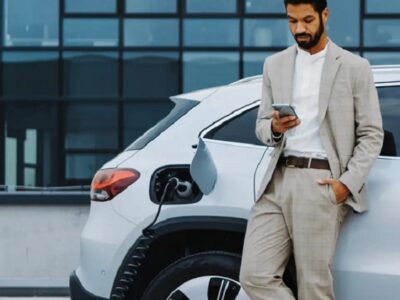 Businessman in a light suit stands beside a white electric SUV, looking at his phone as the charging cable plugged into the car charges the battery near a modern office building.