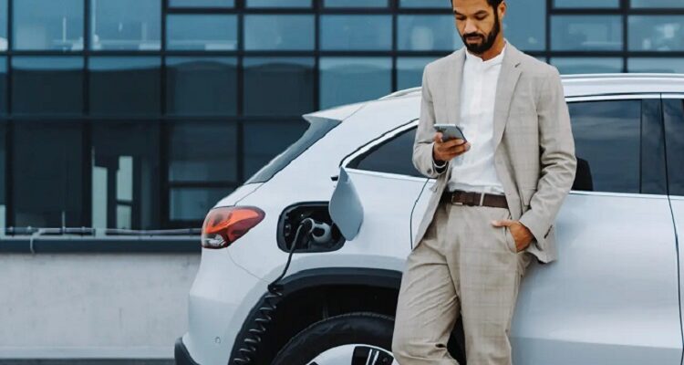Businessman in a light suit stands beside a white electric SUV, looking at his phone as the charging cable plugged into the car charges the battery near a modern office building.